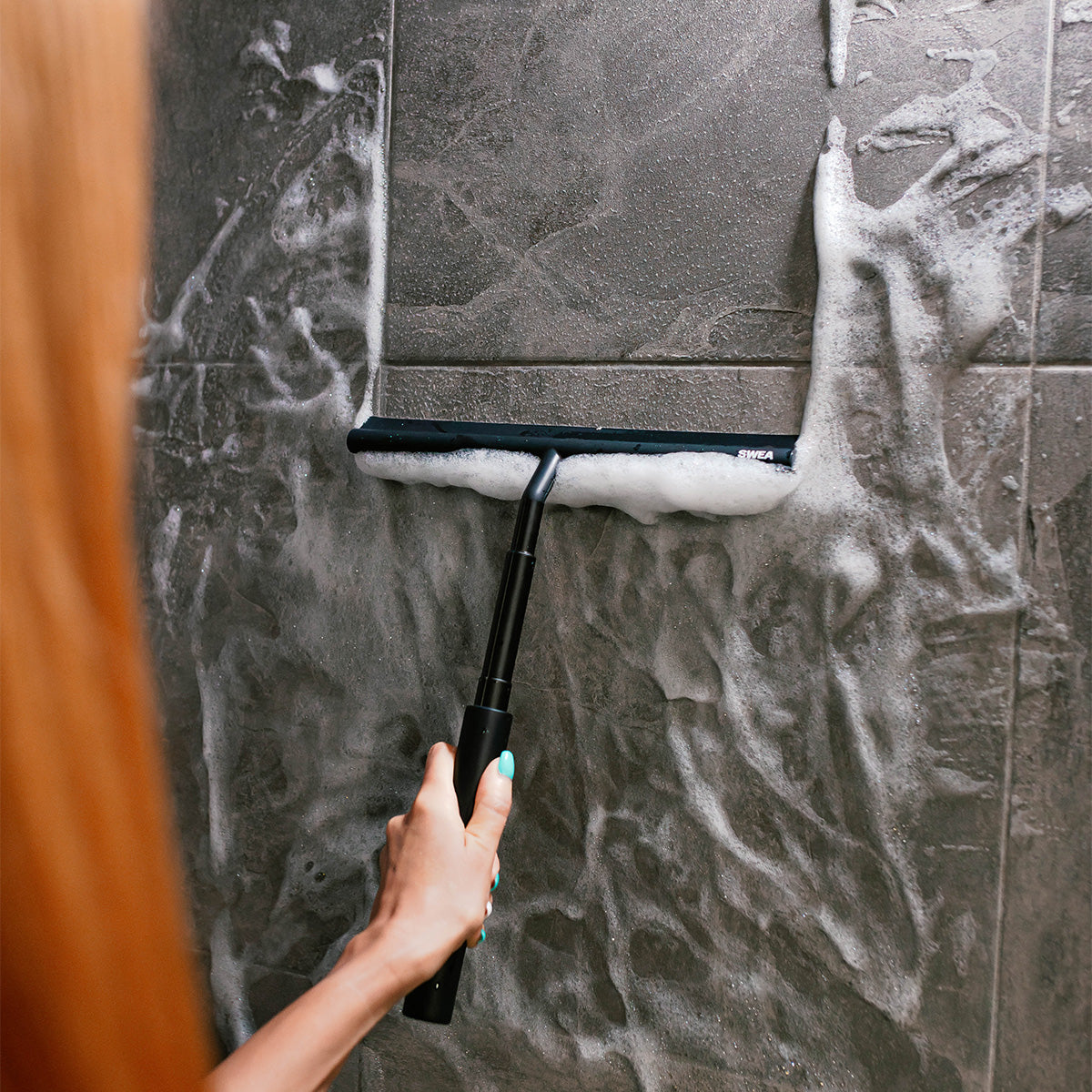 An individual cleaning a shower tiles using the black SWEA squeegee.